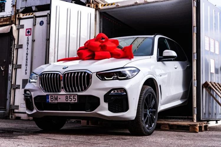 White BMW SUV decorated with a red bow, parked beside shipping containers
