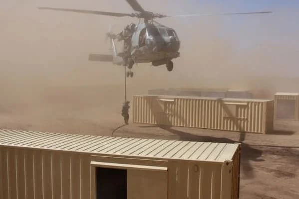 Military helicopter performing a fast-rope insertion onto a row of shipping containers in a desert deployment
